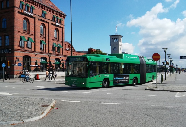 En grön stadsbuss kör genom en öppen korsning med cyklister och gångtrafikanter i närheten. I bakgrunden syns en stor röd tegelbyggnad med gröna markiser, märkt "Restaurang", samt ett vitt klocktorn med blå himmel och vita moln ovanför.