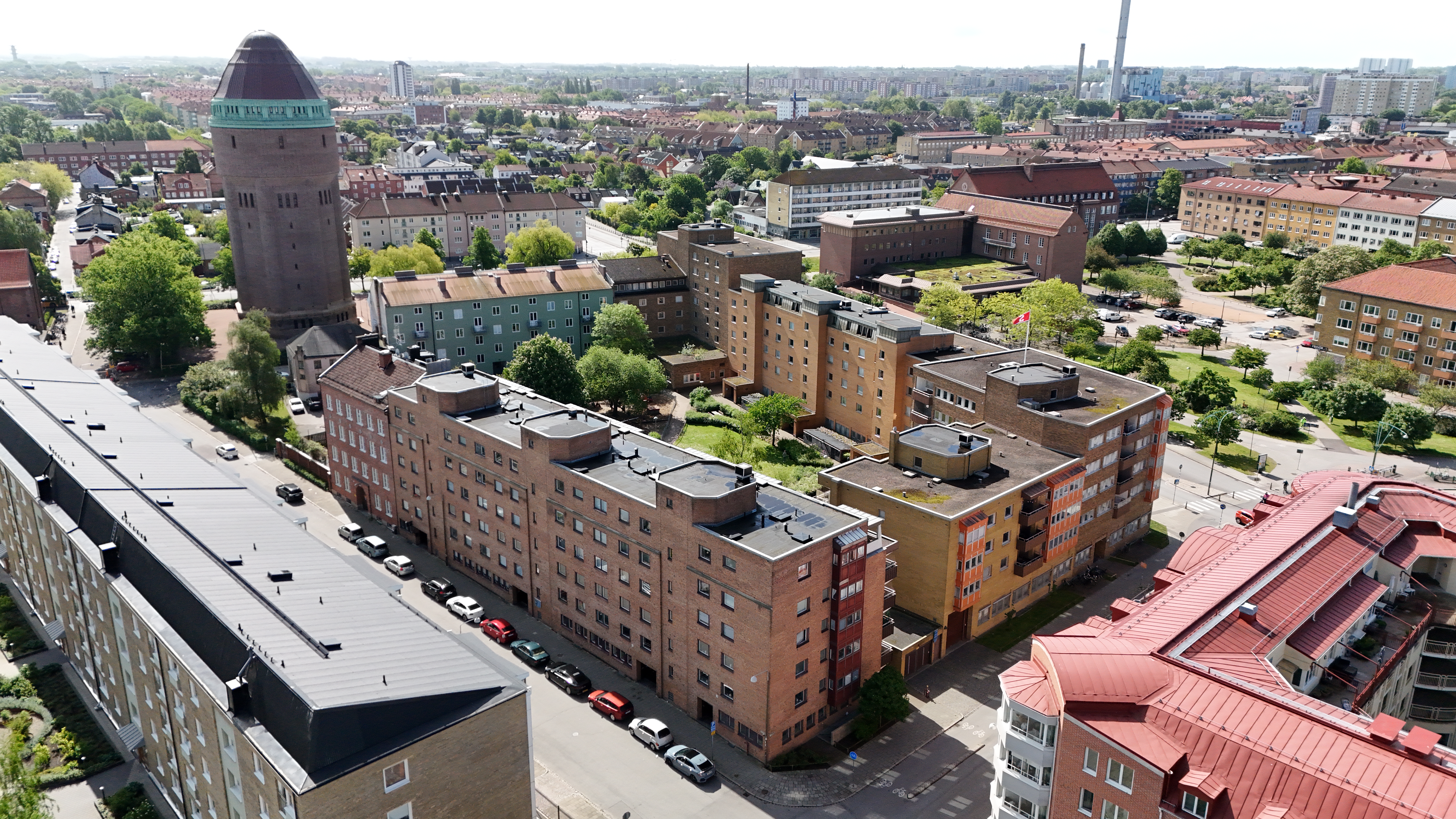 Flygbild över ett bostadsområde i stadsmiljö med flerbostadshus, grönska och en stor vattentornsliknande byggnad i bakgrunden.