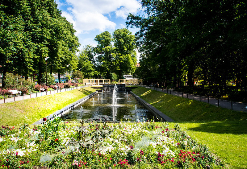 En lummig parkmiljö med en långsmal kanal i mitten, kantad av välklippta grässlänter och prunkande blomrabatter i förgrunden. I kanalen porlar en fontän och längre bort syns en liten bro. Träden runtom skapar en grön vägg och ger parken en inramad, fridfull känsla.