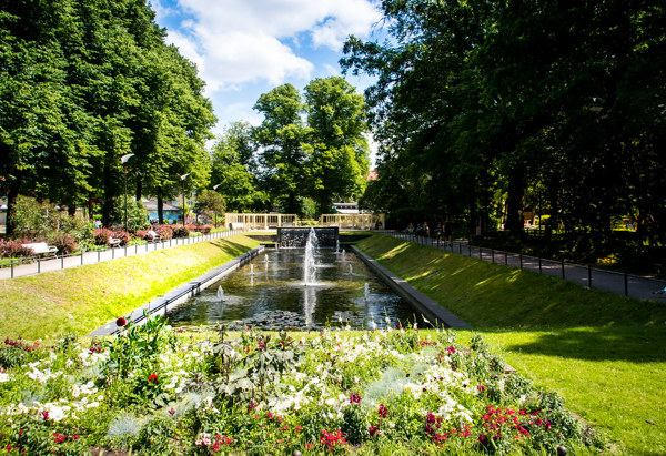 En lummig parkmiljö med en långsmal kanal i mitten, kantad av välklippta grässlänter och prunkande blomrabatter i förgrunden. I kanalen porlar en fontän och längre bort syns en liten bro. Träden runtom skapar en grön vägg och ger parken en inramad, fridfull känsla.
