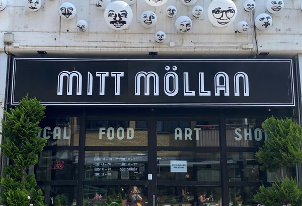 Entrén till Mitt Möllan, ett kreativt centrum för mat, konst och butiker. Ovanför den svarta skylten med vit text sitter dekorativa bollar med ansikten, vilket ger platsen en lekfull och konstnärlig karaktär. Gröna växter i krukor ramar in ingången.
