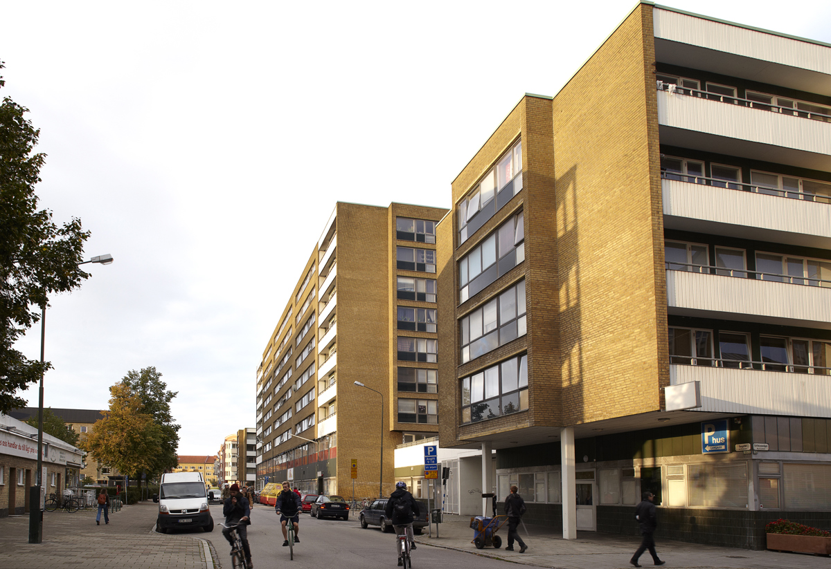 En stadsgata kantad av höga bostadshus i gult tegel med vita balkonger. Flera personer cyklar och promenerar längs trottoaren, och en man ses med en rullator. En vit skåpbil är parkerad vid sidan av vägen och träden visar höstfärger i bakgrunden.