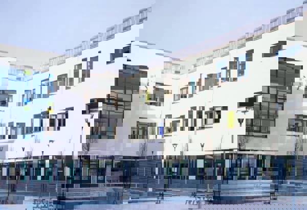 En modern skolbyggnad med vit fasad och stora fönsterpartier i varierande färger. Byggnaden omger en öppen innergård med bänkar, lampstolpar och unga träd. En stor klocka är synlig på ena väggen, och gården är stenbelagd med geometriska mönster.