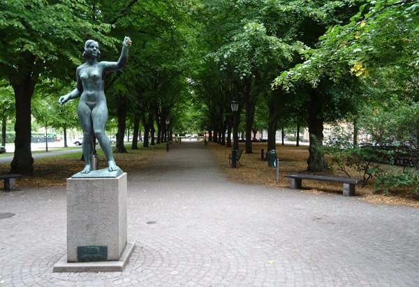 En staty av en naken kvinna i brons står på ett stenfundament i en allé med träd som bildar en tunnel över en gångväg. På båda sidor om gången finns parkbänkar och träd med gröna blad. Marken är täckt av stenplattor och vissna löv.