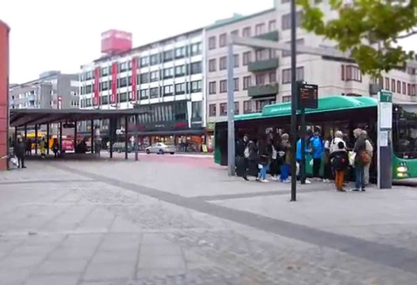 En stadsmiljö med en busshållplats där många människor väntar eller kliver på en grön stadsbuss. I bakgrunden syns fler bussar, flerfamiljshus och affärer. Det är mulet väder och trottoaren är belagd med grå plattor och kullersten.