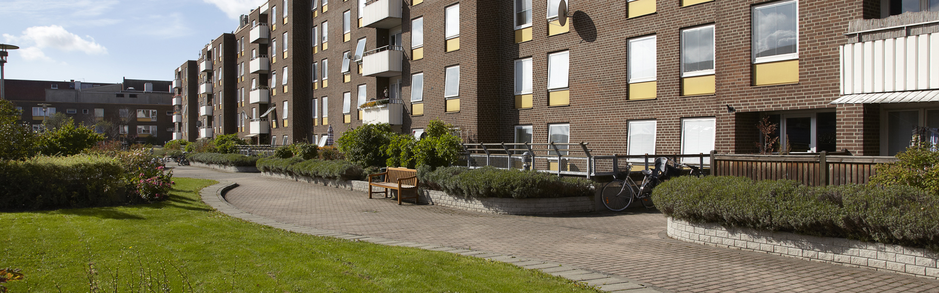 Ett bostadshus i tegel med fyra våningar, typiskt för miljonprogrammets stil. Byggnaden omges av en välskött innergård med grönytor, buskar och gångvägar – en lugn och funktionell miljö för de boende. Balkongerna och de symmetriskt placerade fönstren ger fasaden ett enkelt men rytmiskt uttryck.
