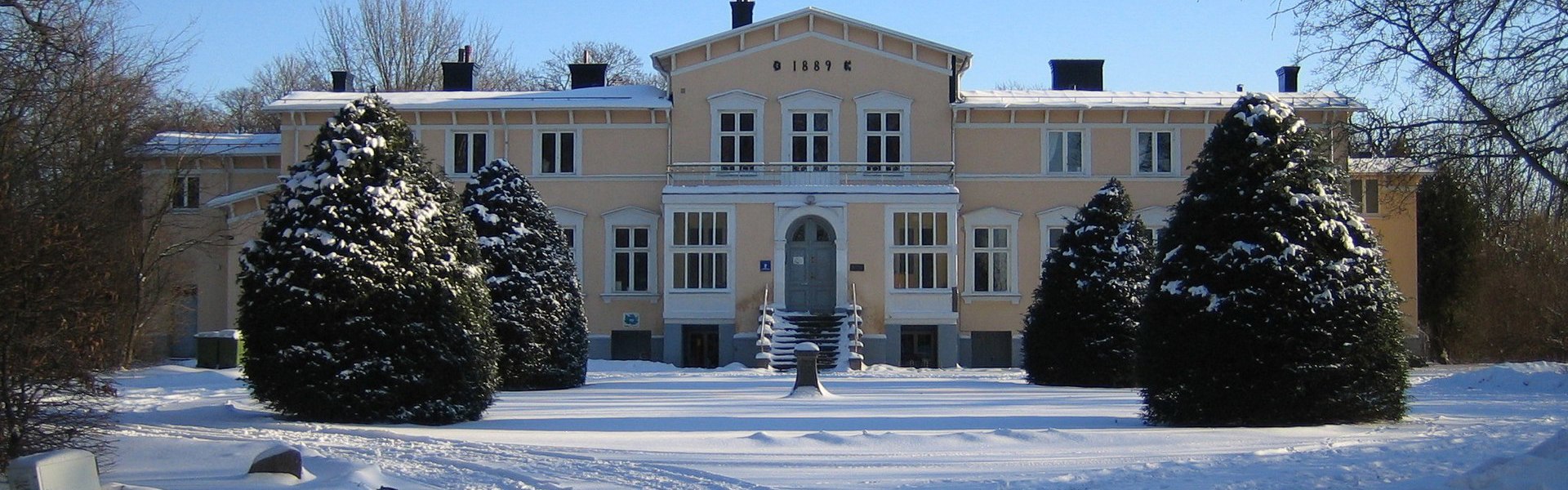 Ett stort, ljust hus i vintermiljö, omgiven av snötäckta granar och ett snötäckt uppfartsspår. Klarblå himmel och vintriga träd ramas in i bakgrunden.