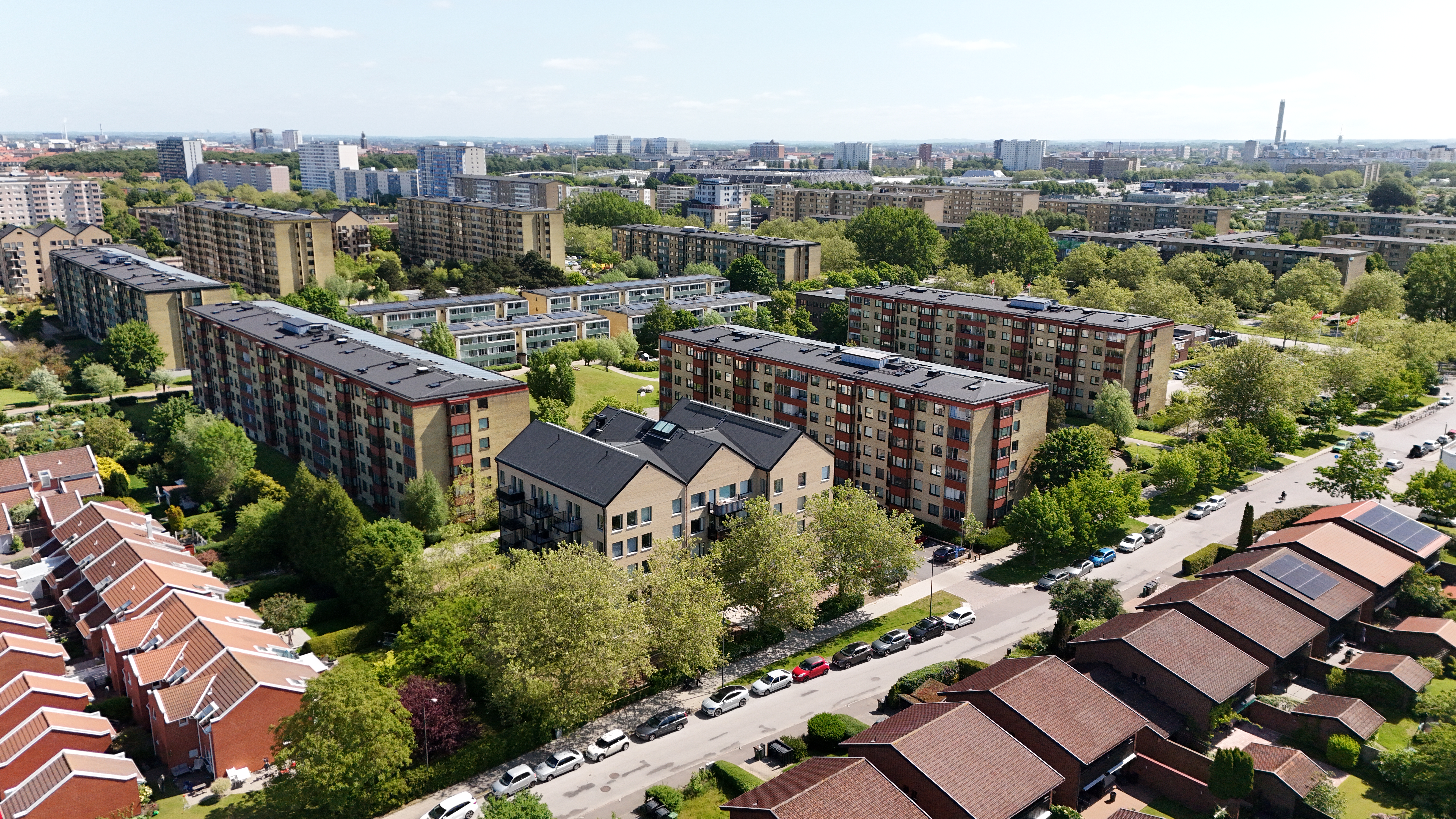 Flygbild över ett grönt bostadsområde med flera flerbostadshus, villor och parkerade bilar längs gatorna.