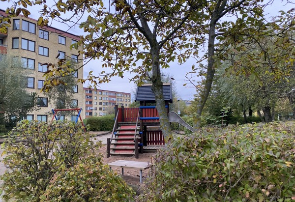 En lekplats med en trälekstruktur som har en rutschkana och trappor, omgiven av buskar och träd. I bakgrunden syns flerbostadshus med färgglada fasader. Höstlöv syns i träden och på marken.