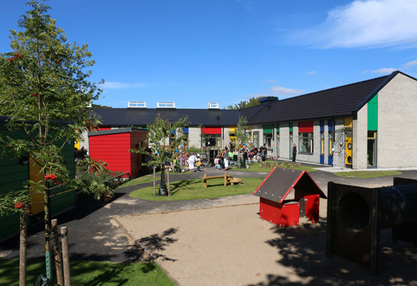 Förskolegård med flera barn som leker utomhus en solig dag. Gården omges av moderna byggnader med färgglada detaljer. Det finns lekstugor, träd och bänkar på området.