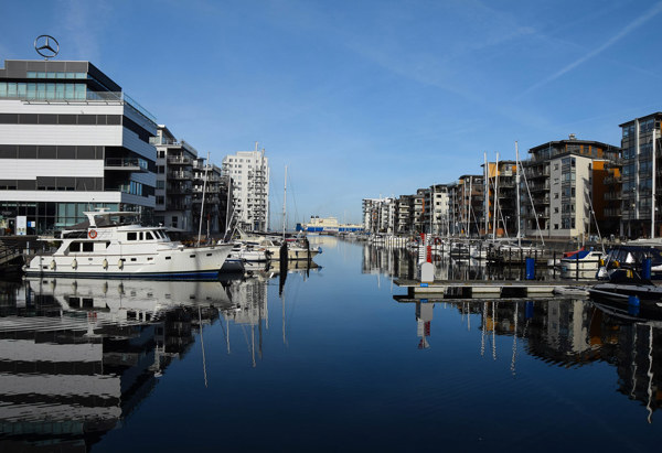 En småbåtshamn med moderna byggnader på båda sidor, båtar förtöjda vid bryggor och en spegelblank vattenyta under en klarblå himmel.