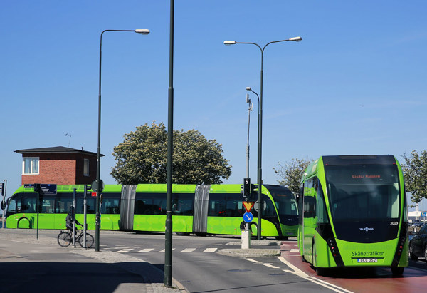 En grön buss kör på en gata under en klarblå himmel, med en cykel parkerad vid trottoaren och träd i bakgrunden.