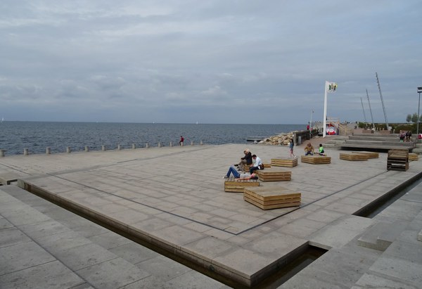 En öppen plats vid havet med sittplatser i trä, människor som kopplar av och en molnig himmel i bakgrunden.