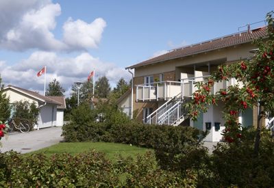 Ett bostadsområde med tvåvåningshus i tegel och balkonger. Framför huset finns buskar med röda bär. Två flaggstänger med hissade flaggor syns i bakgrunden, och en cykel står parkerad vid en byggnad. Himlen är delvis molnig.