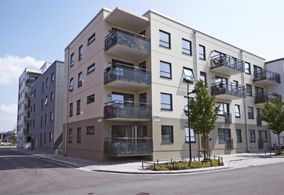 Ett modernt fyravåningshus med balkonger, omgiven av träd och en trottoar i ett bostadsområde.