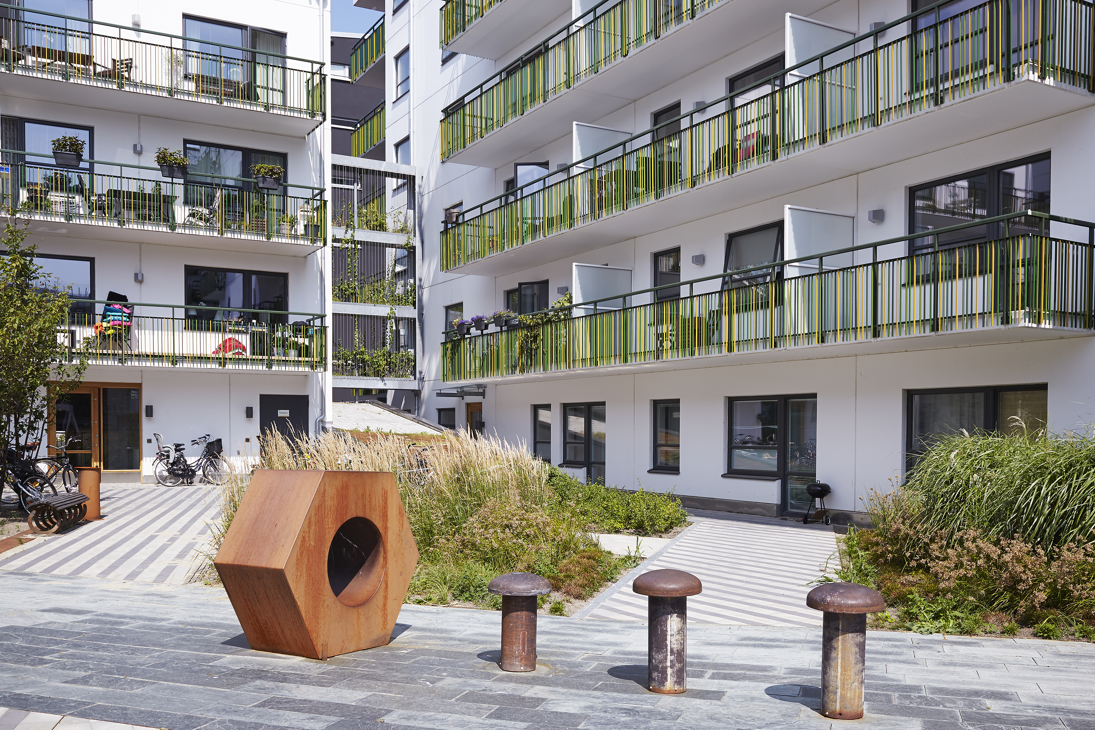 En innergård med moderna lägenhetsbyggnader, gröna balkongräcken, växter och en stor, geometrisk skulptur i förgrunden.
