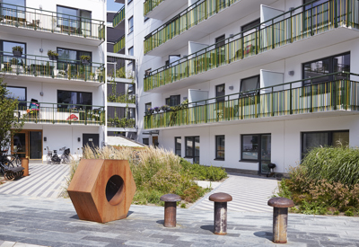 En innergård med moderna lägenhetsbyggnader, gröna balkongräcken, växter och en stor, geometrisk skulptur i förgrunden.
