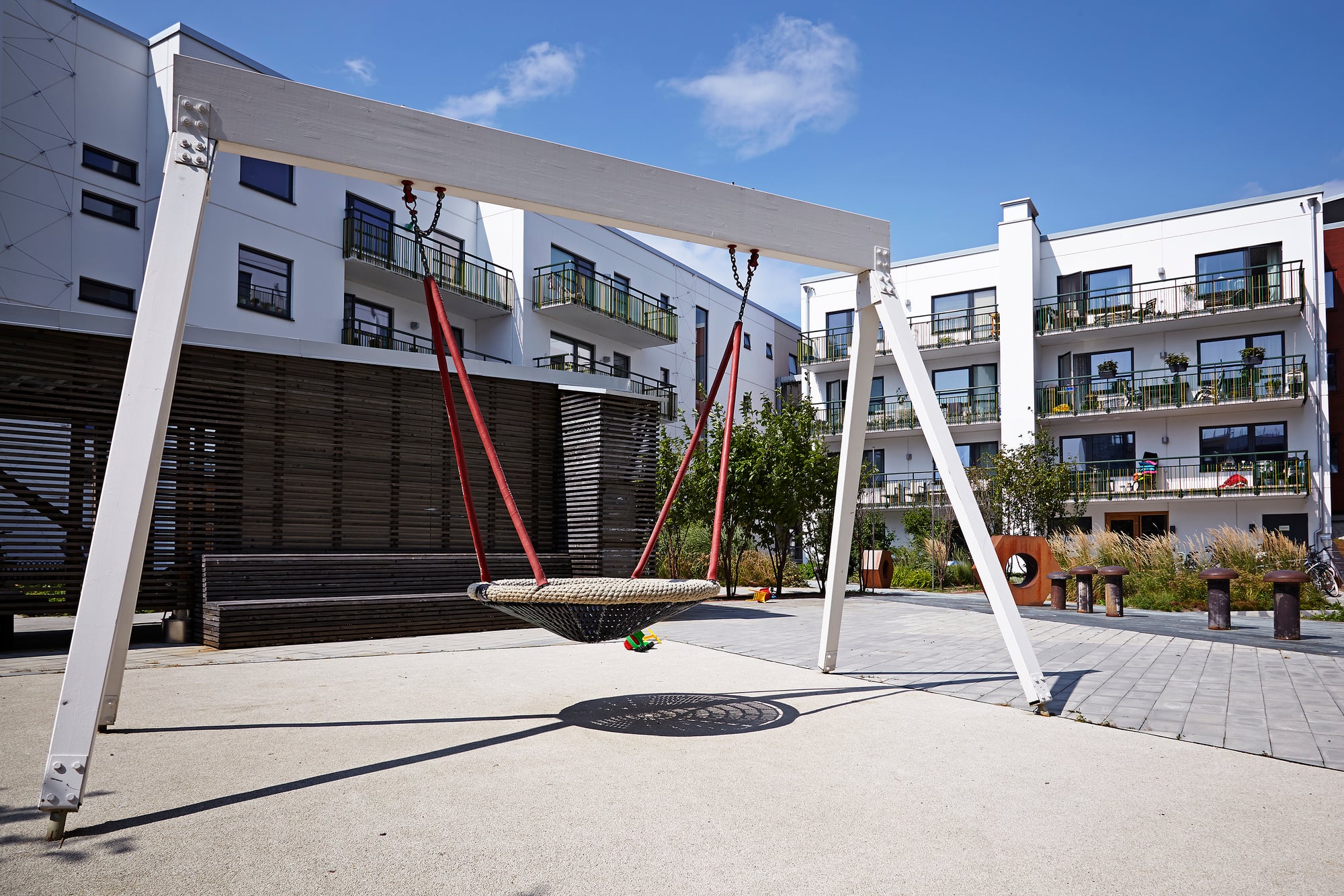 Lekplats på innergård med stor gunga, vita flerbostadshus, balkonger och planteringar i bakgrunden.