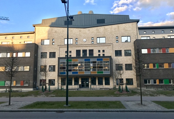 En modern skolbyggnad med färgglada detaljer på fasaden, omgiven av en trottoar och grönska under en blå himmel med några moln.