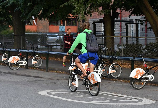 En person cyklar på en hyrcykel i en park med fler cyklar parkerade i bakgrunden, omgiven av träd och trottoarer.
