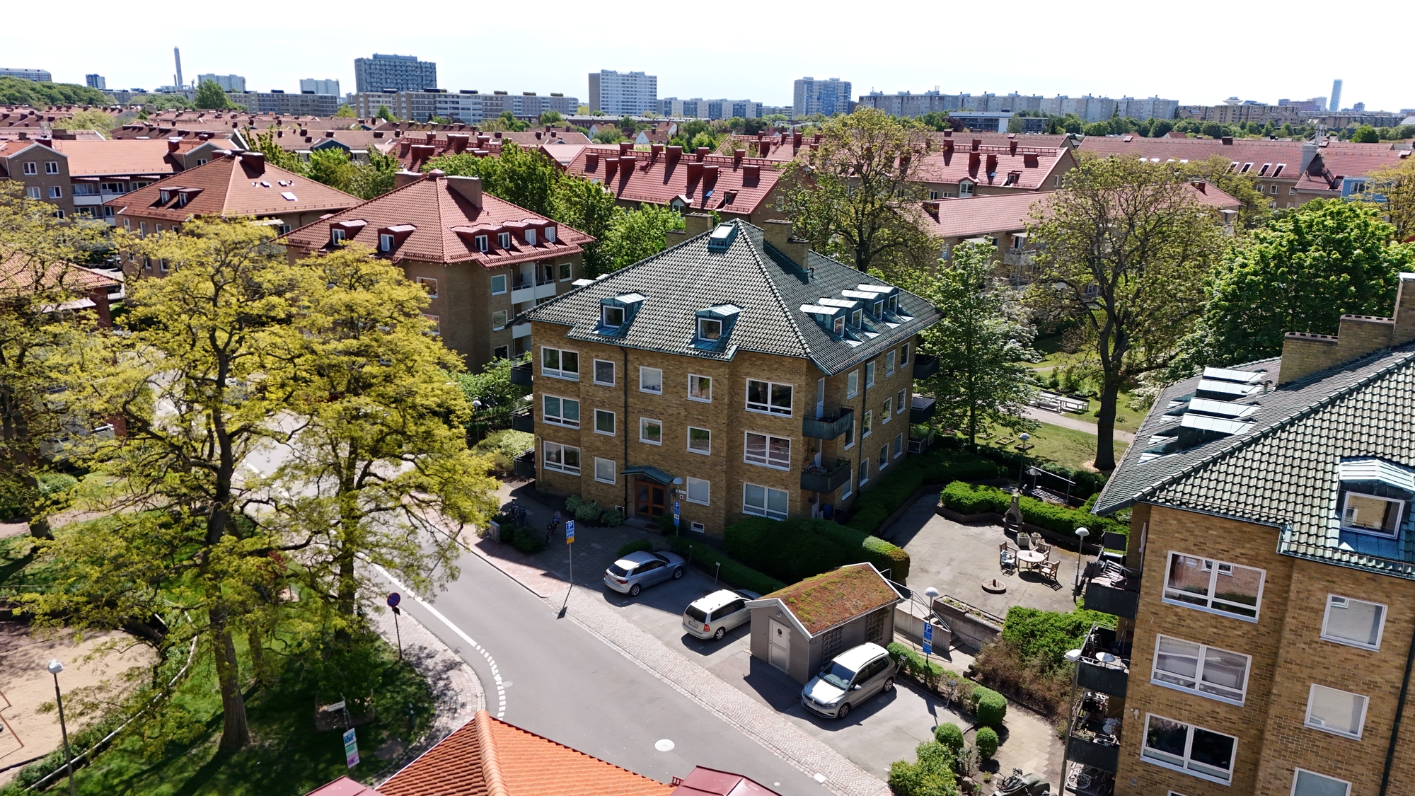 Flygbild över ett bostadsområde med flerbostadshus, gator, parkerade bilar och mycket grönska. I bakgrunden syns fler byggnader och stadssiluett under en klar himmel.