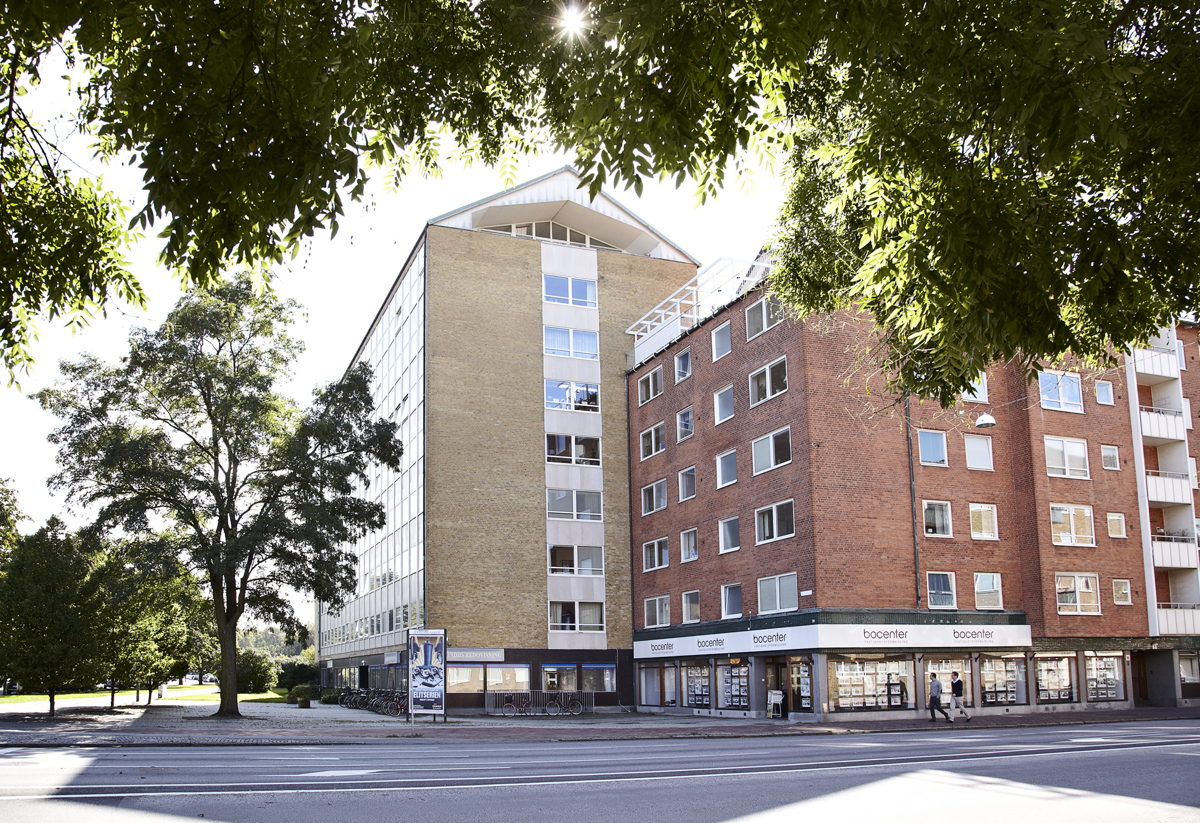 Gatubild med två sammanbyggda flerbostadshus i beige och rött tegel med butikslokaler i bottenvåningen, omslutna av gröna träd i solljus.