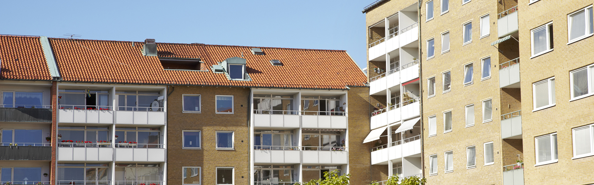 Flerfamiljshus med tegel- och putsfasader, röda tak och balkonger, omgivna av grönska under en klarblå himmel.