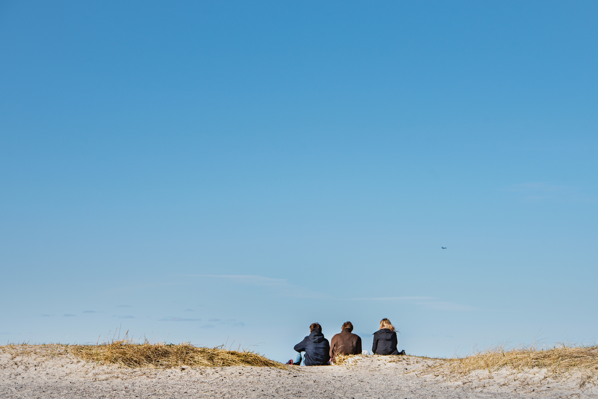 Tre personer sitter på en sanddyn vid stranden och blickar ut mot havet under en klarblå himmel. Bilden har mycket himmel och en lugn, öppen känsla.