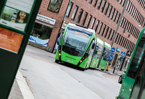 En grön, modern stadsbuss kör genom en stadsgata med röda tegelbyggnader i bakgrunden. I förgrunden syns en del av en busshållplats.