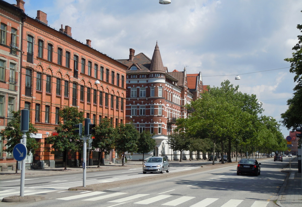 En gatubild i ett urbant område med flera flerfamiljsbyggnader i tegel, träd som kantar trottoaren och några bilar på vägen. Byggnaderna har arkitektoniska detaljer som välvda fönster och dekorativa gesimser. Det finns också gatlyktor och övergångsställen synliga.