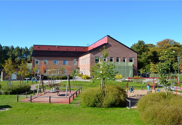 En skolbyggnad med en lekplats i förgrunden. Lekplatsen inkluderar gungor och annan lekutrustning, omgiven av grönt gräs och buskar. Skolbyggnaden är gjord av tegel med stora fönster, och det finns träd runt området.