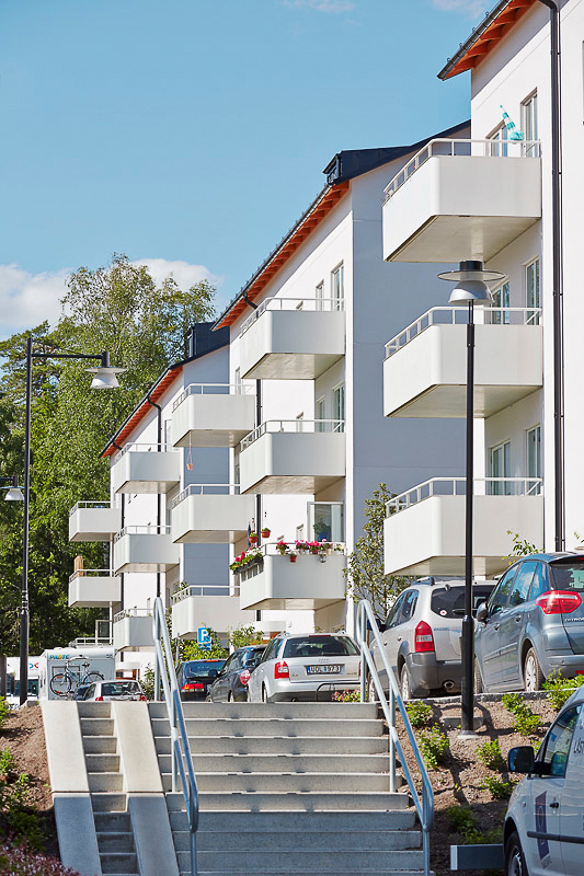 Vita morderna lägenhetshus med vita balkonger. En trapp upp till gatan med parkeringar. Blå himmel och grönska i bakgrunden.
