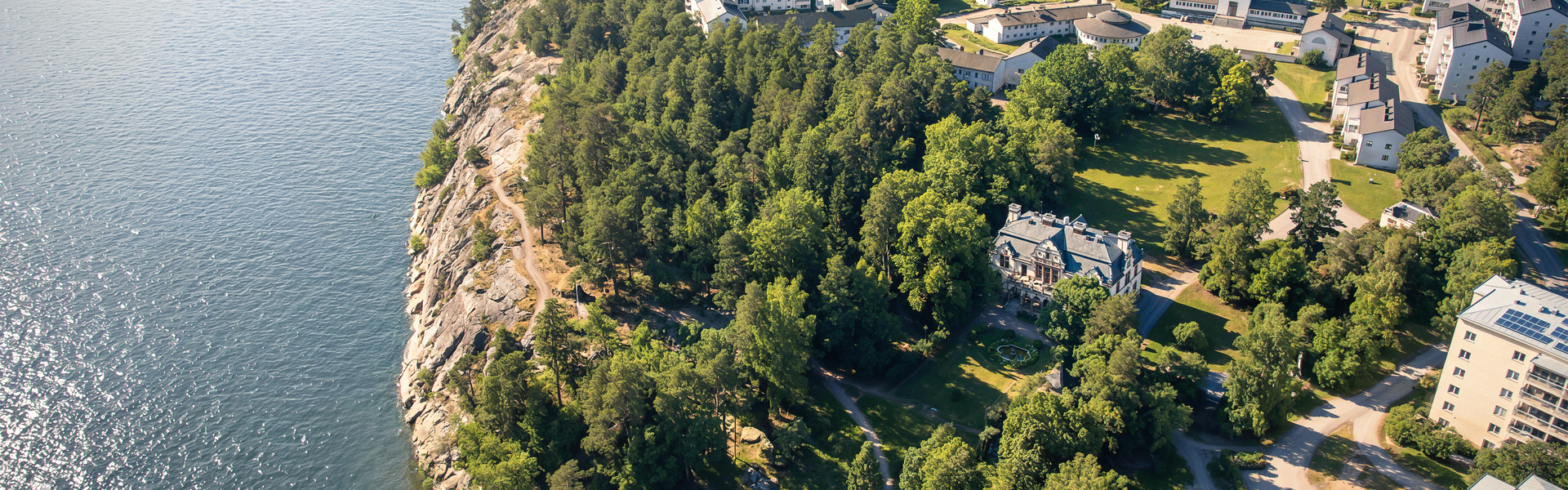 Flygbild över ett grönt bostadsområde vid vattnet, med skog, klippor, gångvägar och flerbostadshus längs strandlinjen.