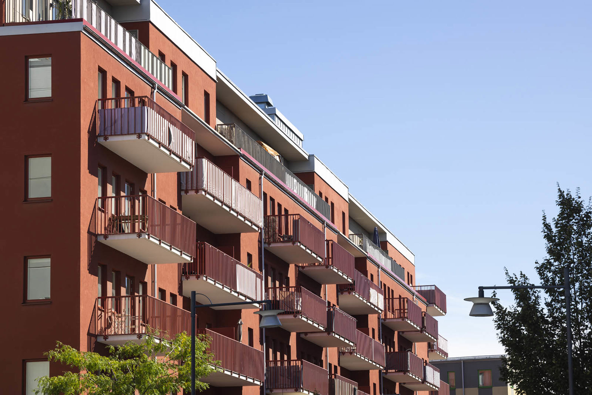 Rött flerbostadshus med flera balkonger i rad under klarblå himmel, med grönska och en lyktstolpe i förgrunden.