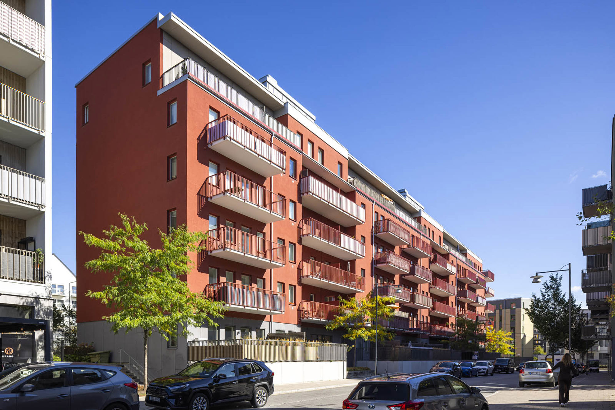 Rött flerbostadshus med många balkonger längs en gata med parkerade bilar, träd och klarblå himmel.