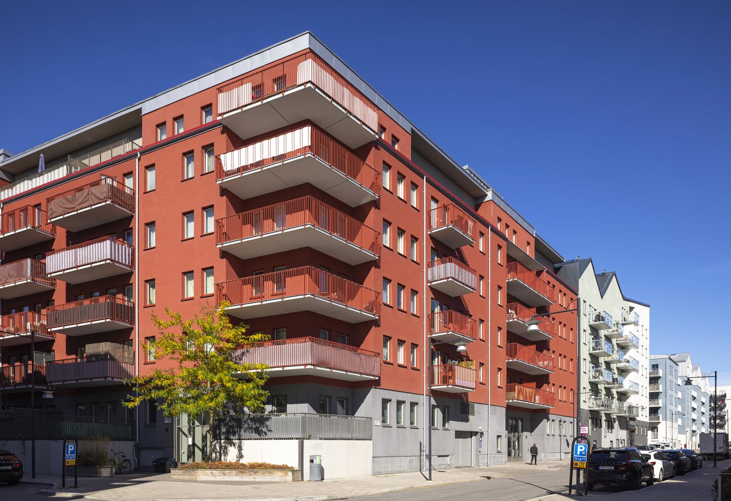 Rött flerfamiljshus med flera våningar och stora balkonger längs fasaden. Byggnaden ligger i ett modernt bostadsområde med bred gata, parkerade bilar och närliggande flerbostadshus i ljusa färger