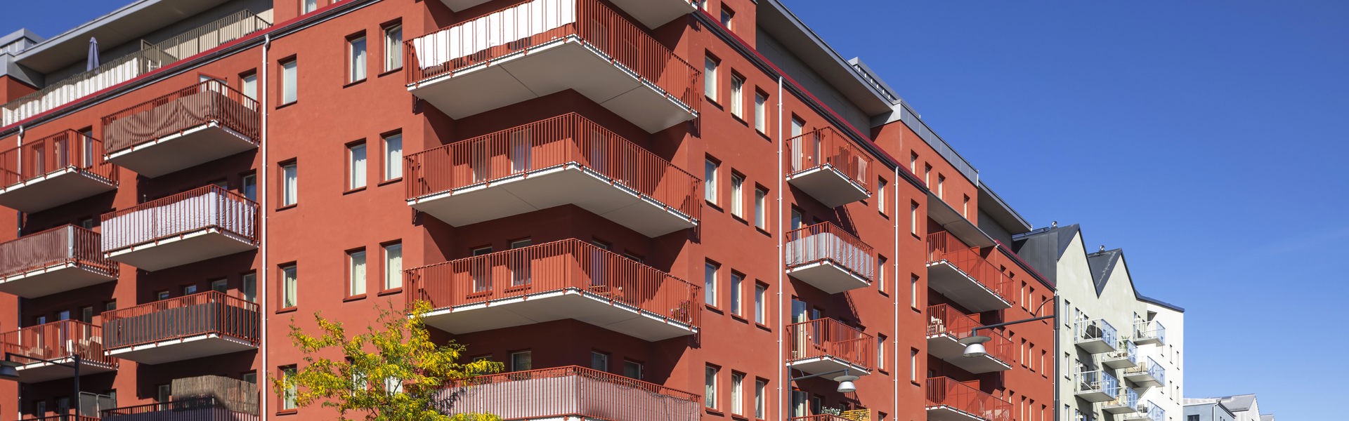 Rött flerfamiljshus med flera våningar och stora balkonger längs fasaden. Byggnaden ligger i ett modernt bostadsområde med bred gata, parkerade bilar och närliggande flerbostadshus i ljusa färger
