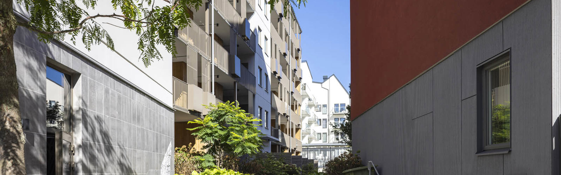 Gångstråk med trappor och grönska mellan flerbostadshus, med röd fasad till höger och ljusa byggnader i bakgrunden.