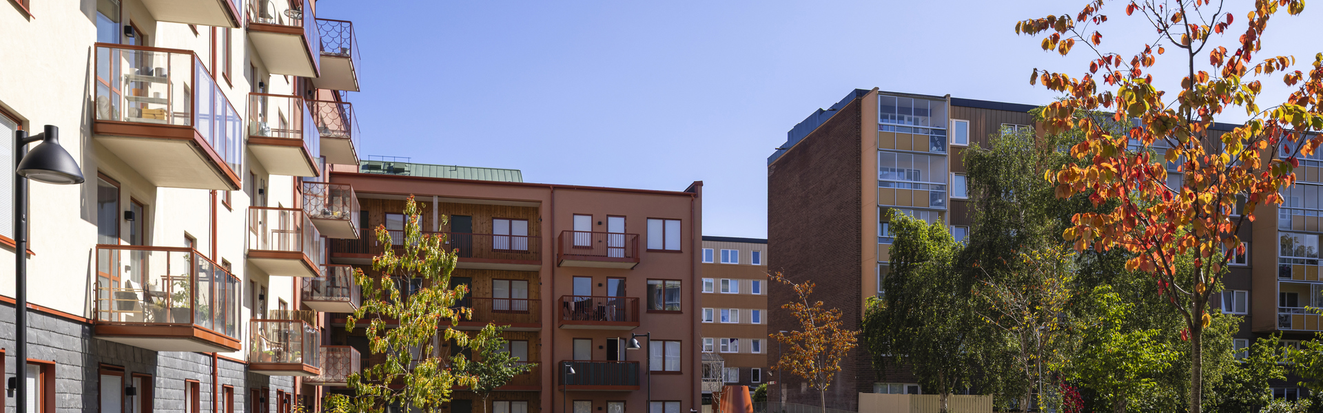 Ett bostadsområde med flerfamiljshus runt en grön innergård med gångvägar, planteringar, bänkar och träd i höstfärger under klarblå himmel.
