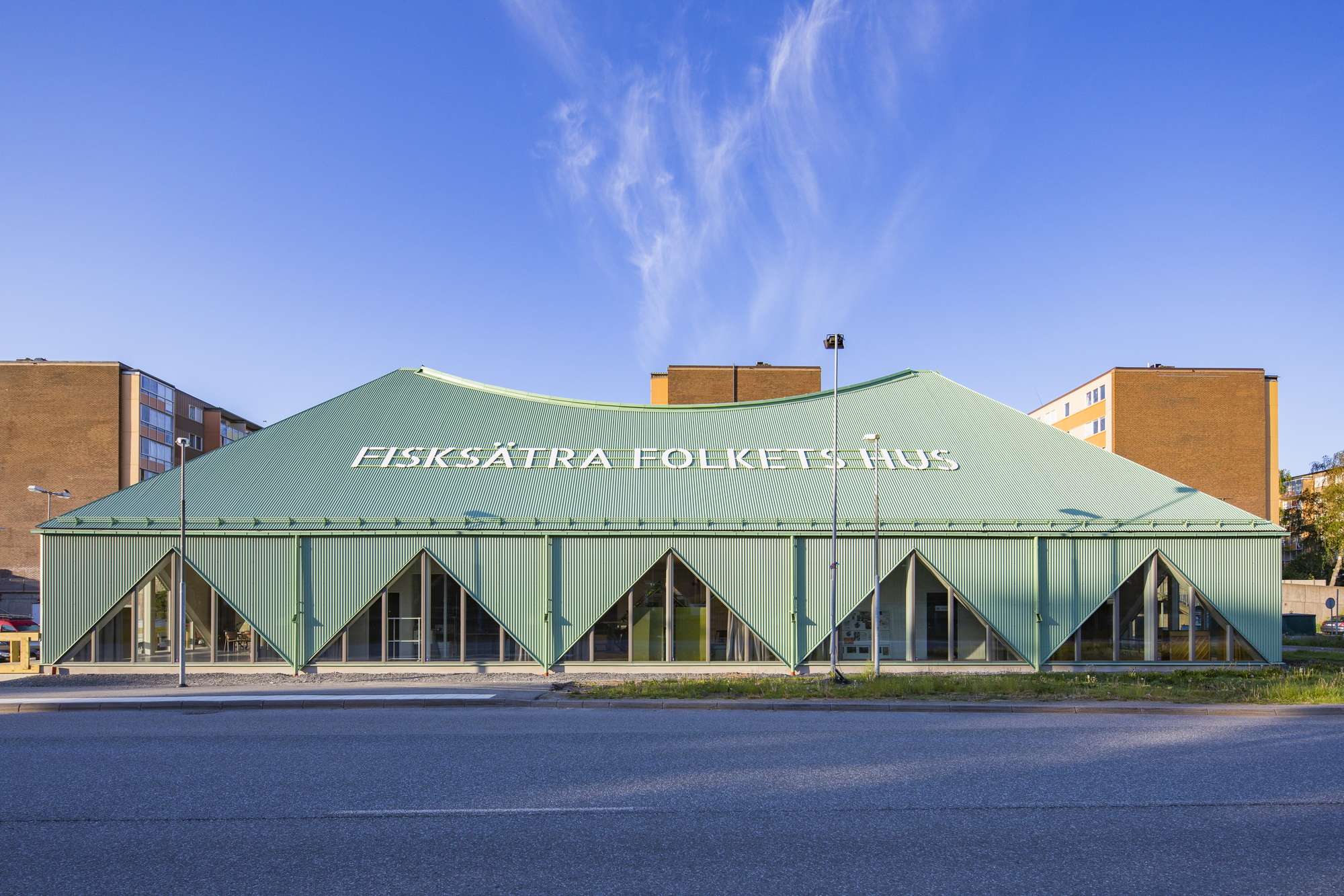 Fasadbild av Fisksätra Folkets Hus med grönt tak och stora fönsterpartier, fotograferad från gatan en solig dag.