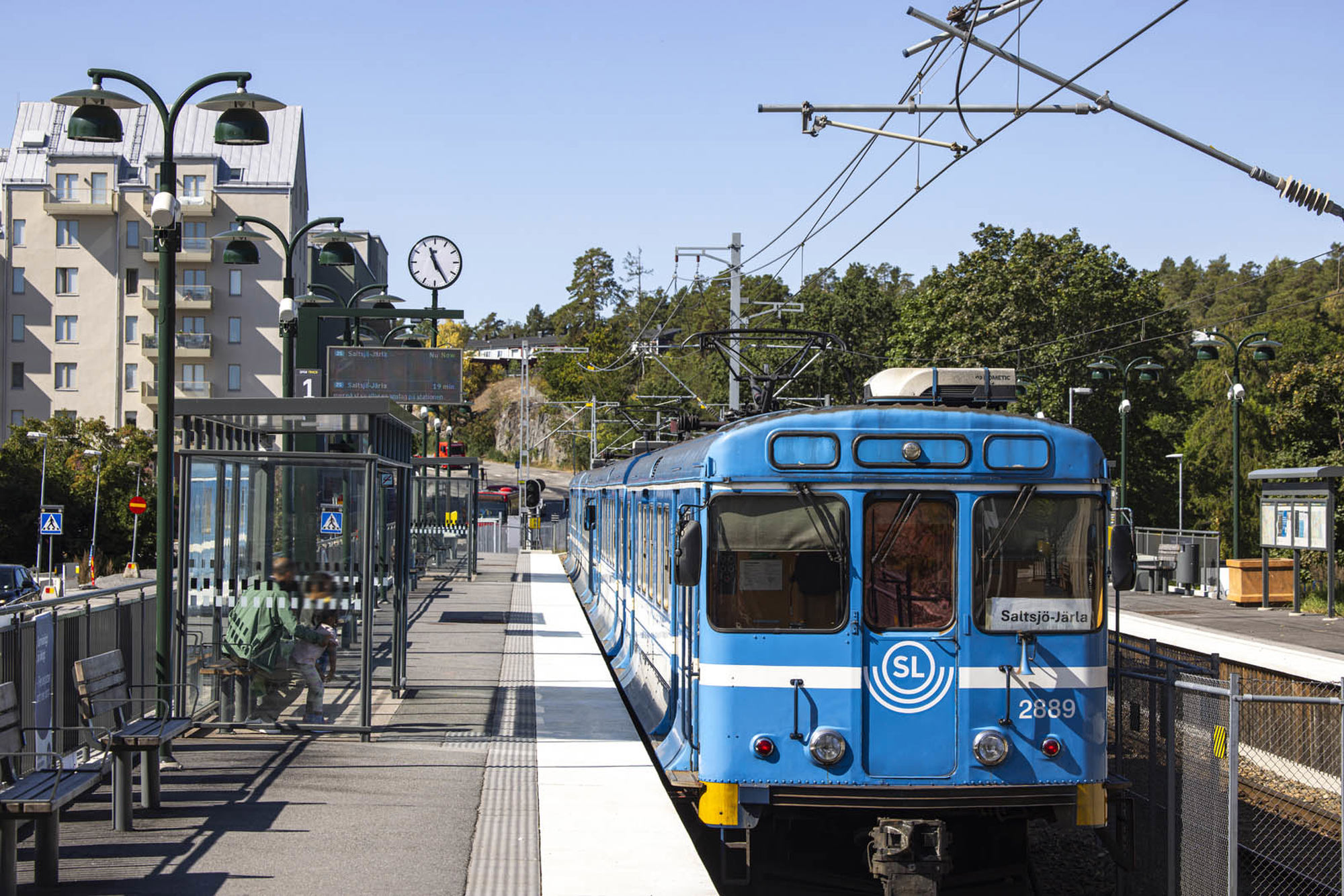 Saltsjöbanans blå tåg vid Fisksätra station. Perrong med kur är person sitter och väntar.Flerfamiljshus i bakgrund