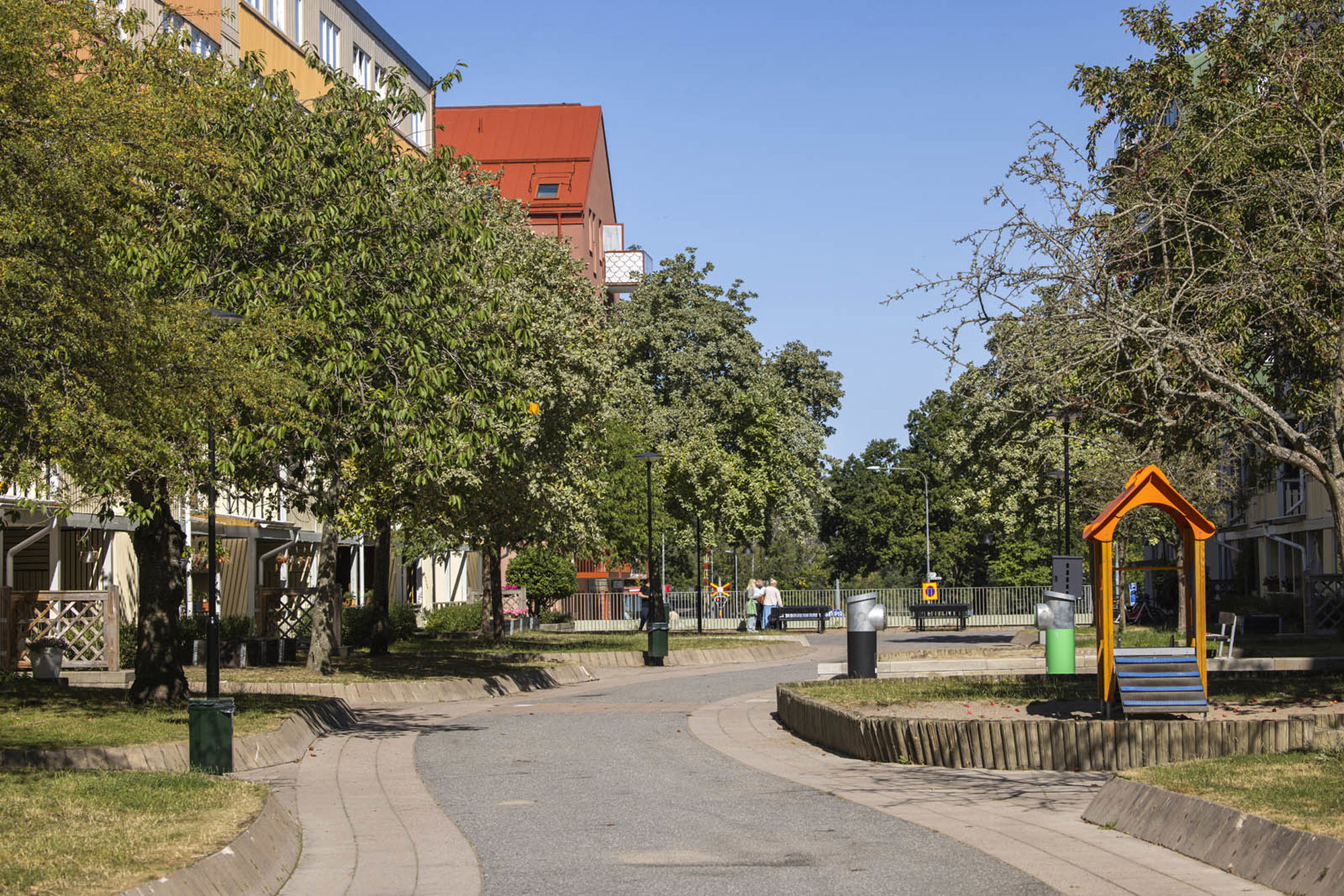 Grönskande gågata mellan flerfamiljshus, träd på vardera sida och en liten lekplats i mitten. 