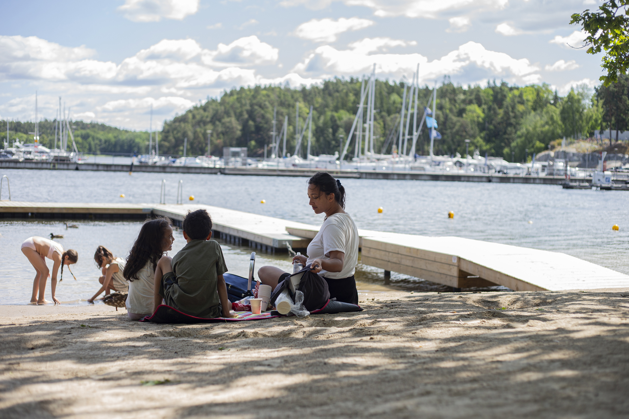 En kvinna med 4 st barn som sitter och leker på en sandstrand med en brygga och en marina i bakgrund. 