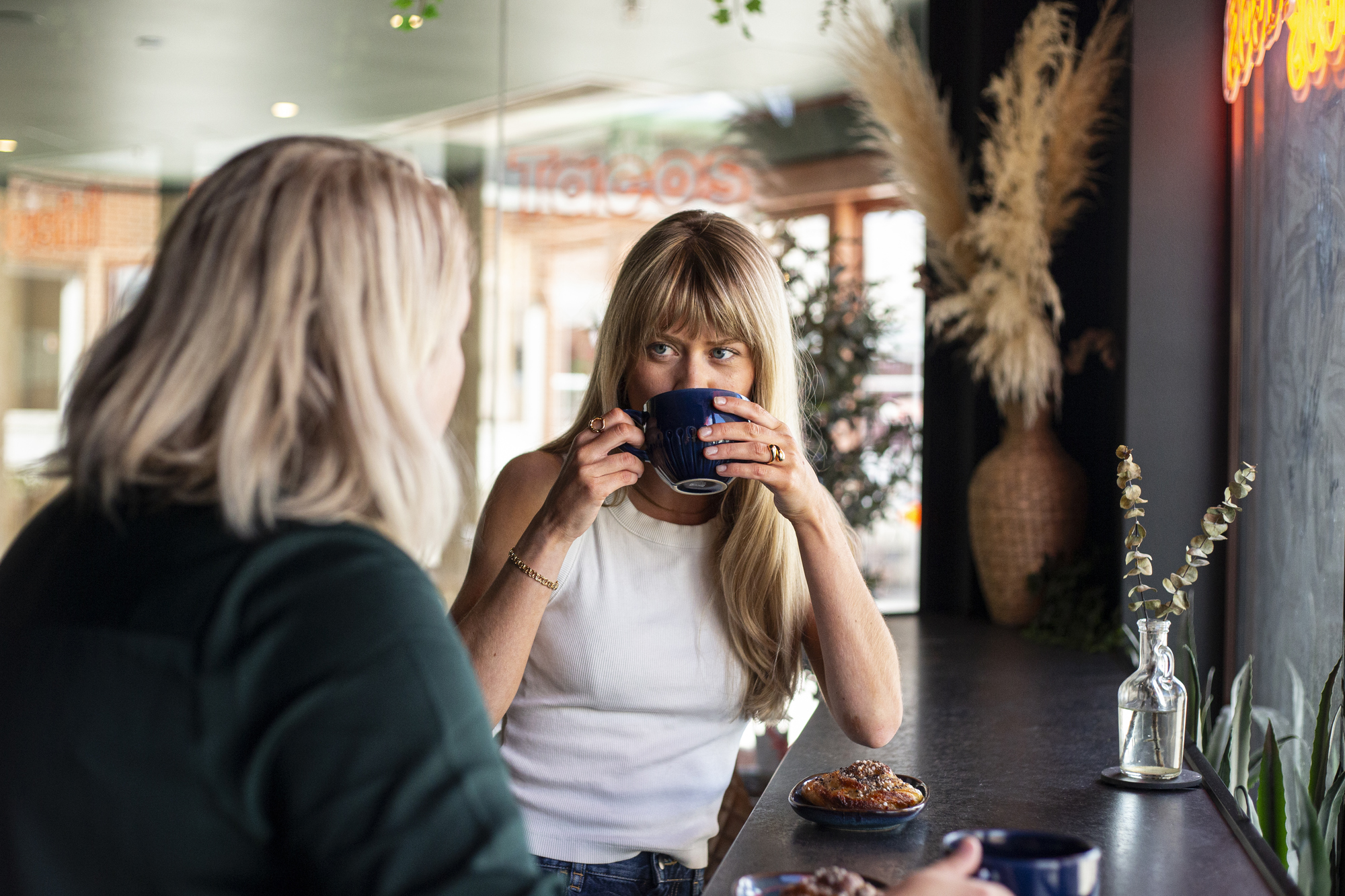 Två kvinnor som sitter ett högre bord och dricker kaffe och äter bullar.