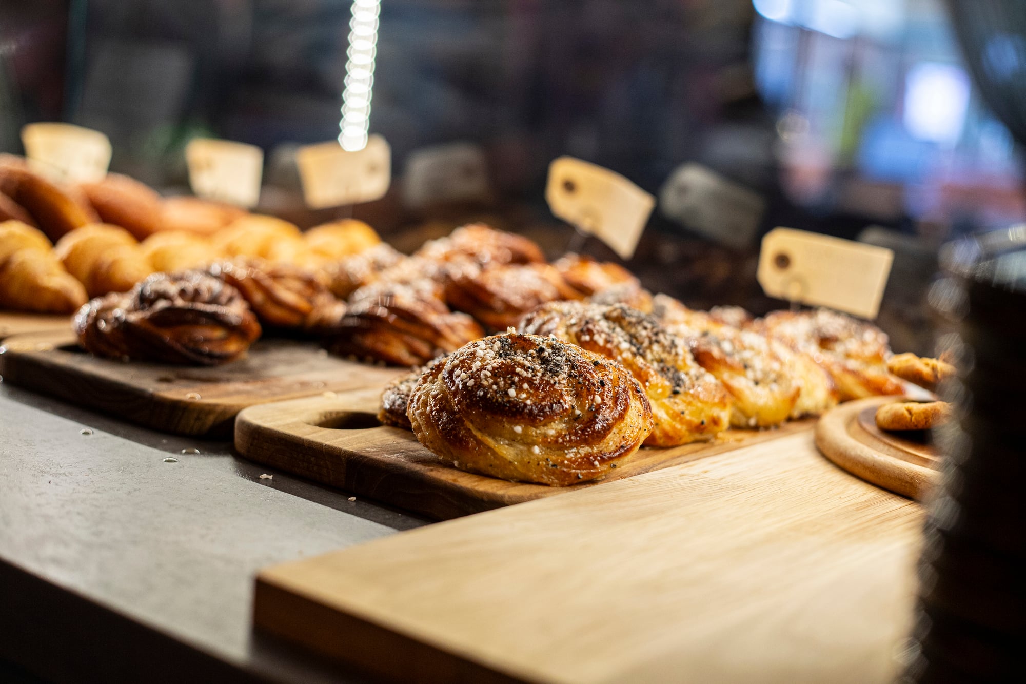  Närbild av nybakade bullar och bakverk på skärbrädor i ett café, med prislappar och suddig bakgrund.