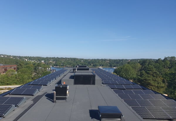 Bild på ett tak med solceller på i en solig miljö med grönska och vatten i bakgrunden.