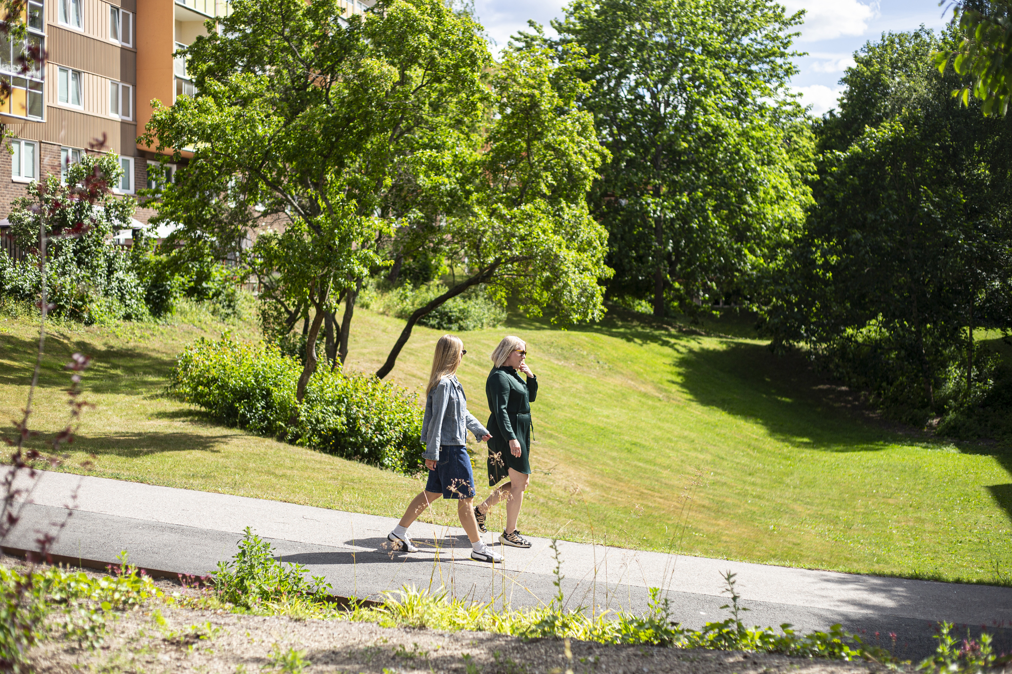Två personer promenerar på en gångväg genom en grön parkmiljö vid bostadshus.