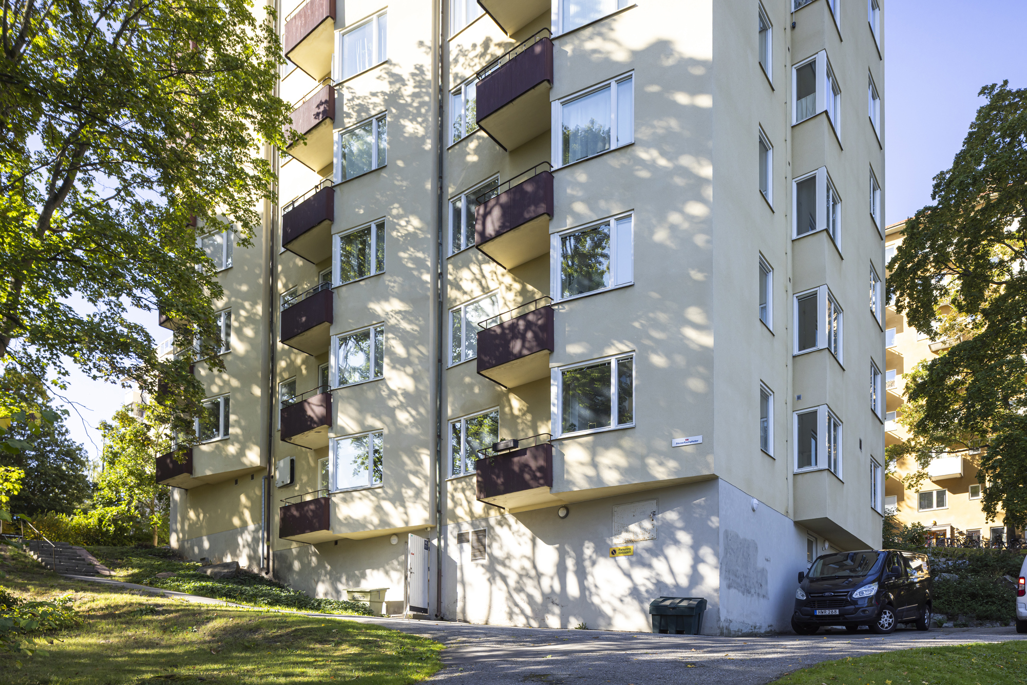 Flerbostadshus med ljus fasad och balkonger, omgivet av grönska och träd.