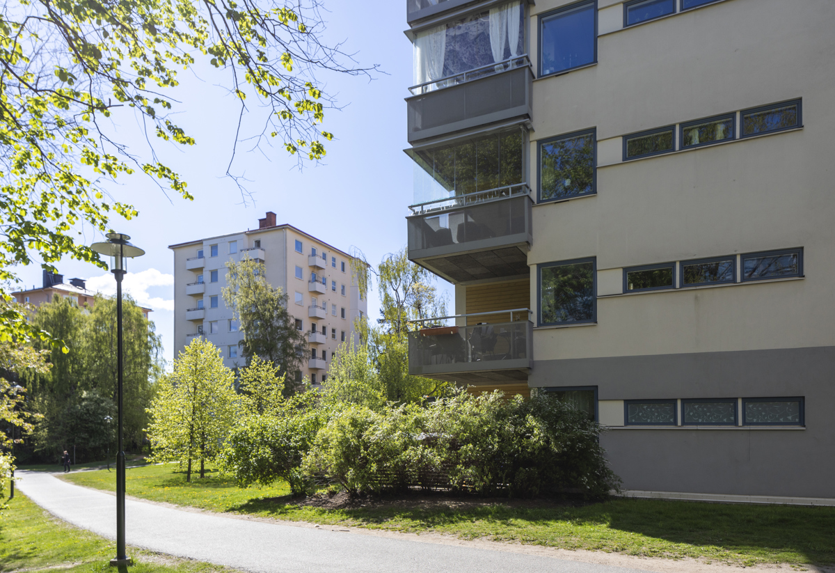 Grenar i förgrunden till vänster, ljusgult flerfamiljshus till höger och ett flerfamiljshus i bakgrunden.