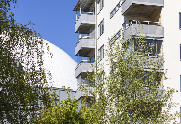 Vitt flerfamiljshus med grå balkonger, grönskande träd i förgrunden och Globen i bakgrunden.